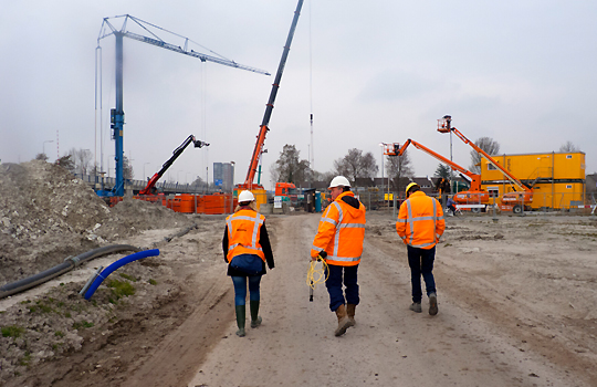 3 bouwvakkers met oranje jassen en een helm lopen op een bouwplaats waar hijskranen staan.
