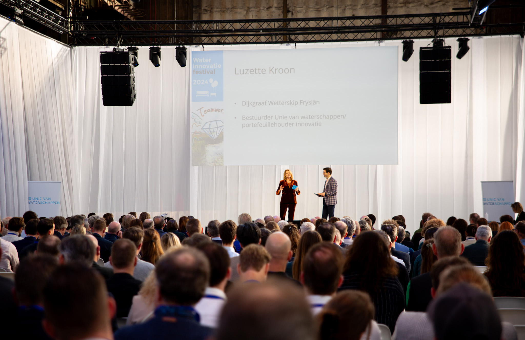 Waterinnovatiefestival: foto van achterin zaal met op podium moderator en Luzette Kroon