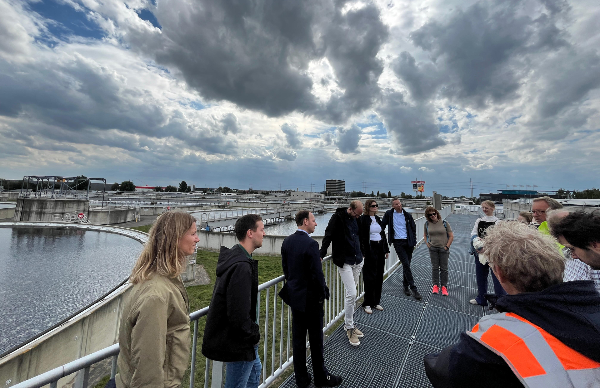 Vertegenwoordigers van de Raad van State staan op een metalen loopbrug van rioolwaterzuiveringsinstallatie Harnaschpolder met uitzicht op de bassins, terwijl een medewerker uitleg geeft. De lucht is deels bewolkt.