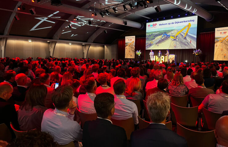 Grote zaal vol publiek tijdens de Dijkwerkersdag 2025 in ’s-Hertogenbosch. Op het podium staan drie sprekers voor een scherm met de tekst “Welkom op de Dijkwerkersdag” en een afbeelding van dijkversterkingswerkzaamheden. De zaal is sfeervol verlicht in rood, en het evenement wordt georganiseerd door het Hoogwaterbeschermingsprogramma (HWBP).