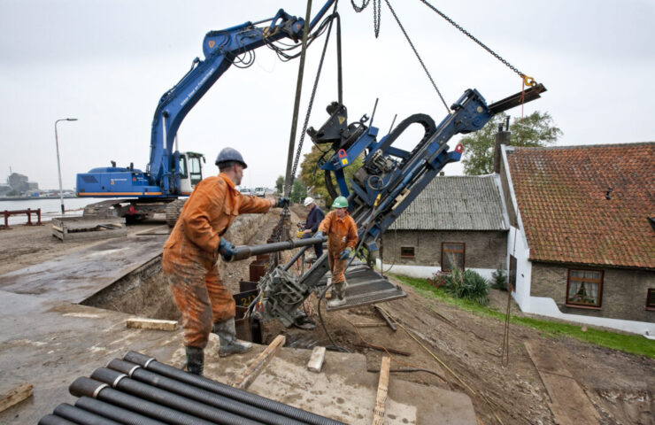 2 werkmannen in oranje overals staan op een dijk naast een hijskraan om elkaar een buis aan te geven.