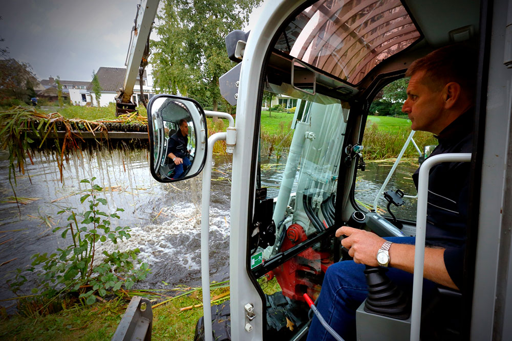 Een man bestuurt een graafmachine langs een watergang. Door het raam en in de zijspiegel is te zien hoe een maaikorf uit het water wordt getild met maaisel. Op de achtergrond staan huizen en bomen.