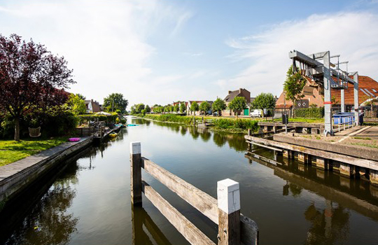 Foto gebruikt voor dec over van het rapport "Monitor implementatie Wet open overheid". De foto toont een waterrijk landschap met woningen, bomen en een brug.