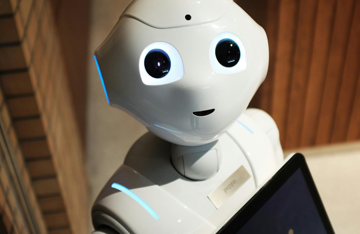 Witte robot met oplichtende ogen kijkt recht in de lens. Robot houdt laptop vast. De achtergrond bestaat uit houten en bakstenen muren