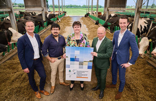 Vertegenwoordigers van LTO Nederland, NAJK, IPO, VNG en de Unie van Waterschappen poseren in een koeienstal met melkkoeien op de achtergrond. Ze houden gezamenlijk een groot bord vast met hun stikstofplant. De omgeving is open en landelijk, met groene weilanden zichtbaar in de verte.