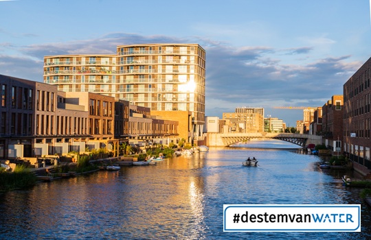 Zicht op een stedelijke gracht met moderne appartementen en woonhuizen langs het water, waar een roeiboot vaart. Op de voorgrond staat de campagnehashtag #destemvanwater in beeld.