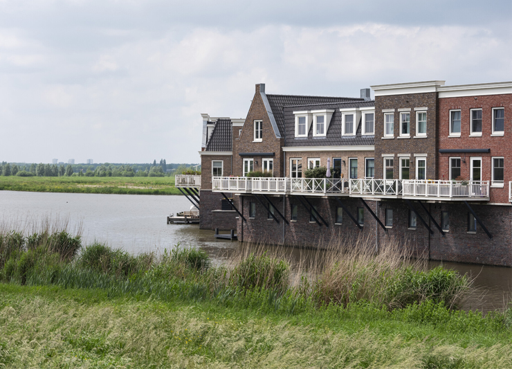 Huizen aan het water. Maar met groot hoogte verschil tussen het water en de woonlaag. Op de voorgrond gras van een dijk.