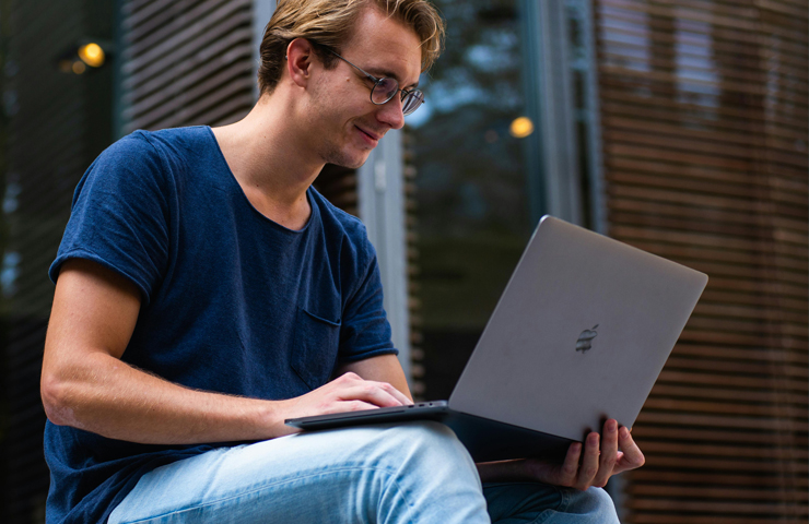 Persoon met laptop buiten voor gebouw met glazen ramen en houten lamellen.