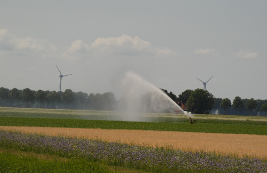 Beregening van akker om gewassen te beschermen tegen droogte en watertekort, met windmolens op de achtergrond.