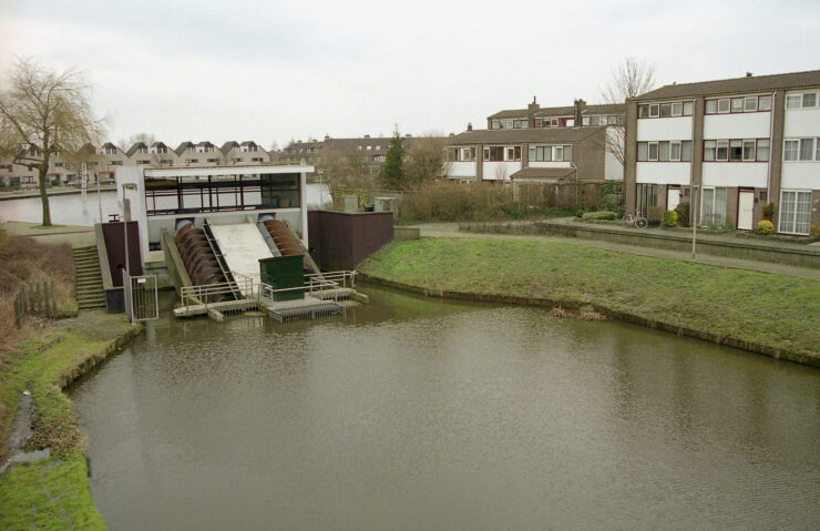 Gemaal met twee grote vijzels in een woonwijk, omringd door watergangen en rijtjeshuizen, als symbool voor het Nederlandse waterbeleid dat centraal staat in het commissiedebat Water.