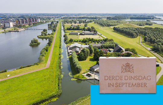 Prinsjesdag de koffer 'Derde Dinsdag in September' tegen een achtergrond van een Nederlands landschap met een stad aan een rivier, een dijk en veel groen.