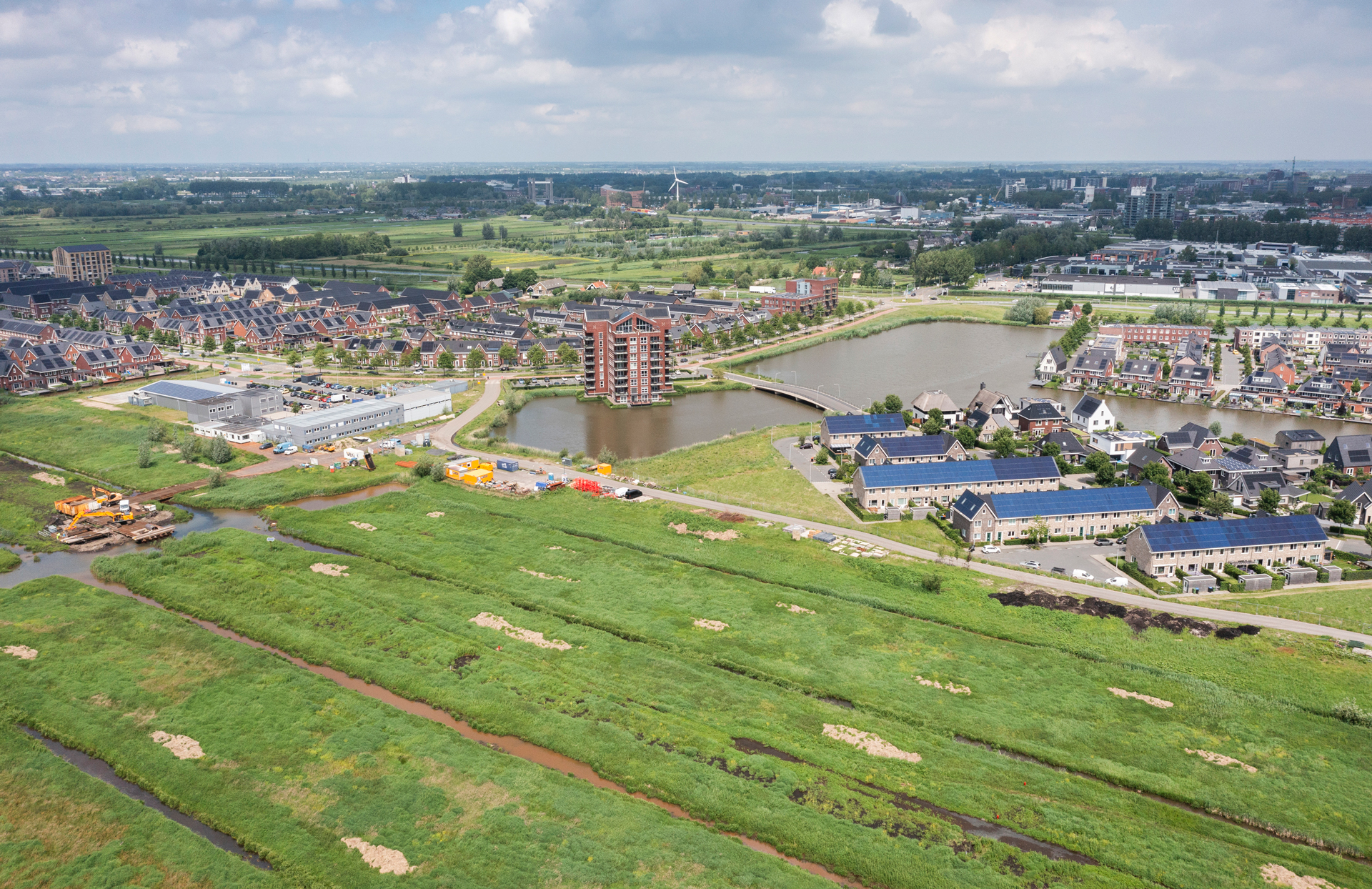 Luchtfoto van een nieuwbouwwijk aan de rand van een stad, omringd door water, groen en veenweidegebied. In het midden een appartementengebouw aan een vijver met brug, op de voorgrond weilanden met sloten en werkzaamheden aan de waterstructuur. De omgeving laat de combinatie van stedelijke ontwikkeling en klimaatadaptieve inrichting zien.