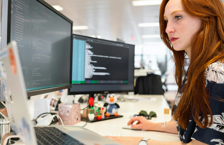 Een jonge vrouw werkt aan computer met twee schermen vol code in een kantooromgeving